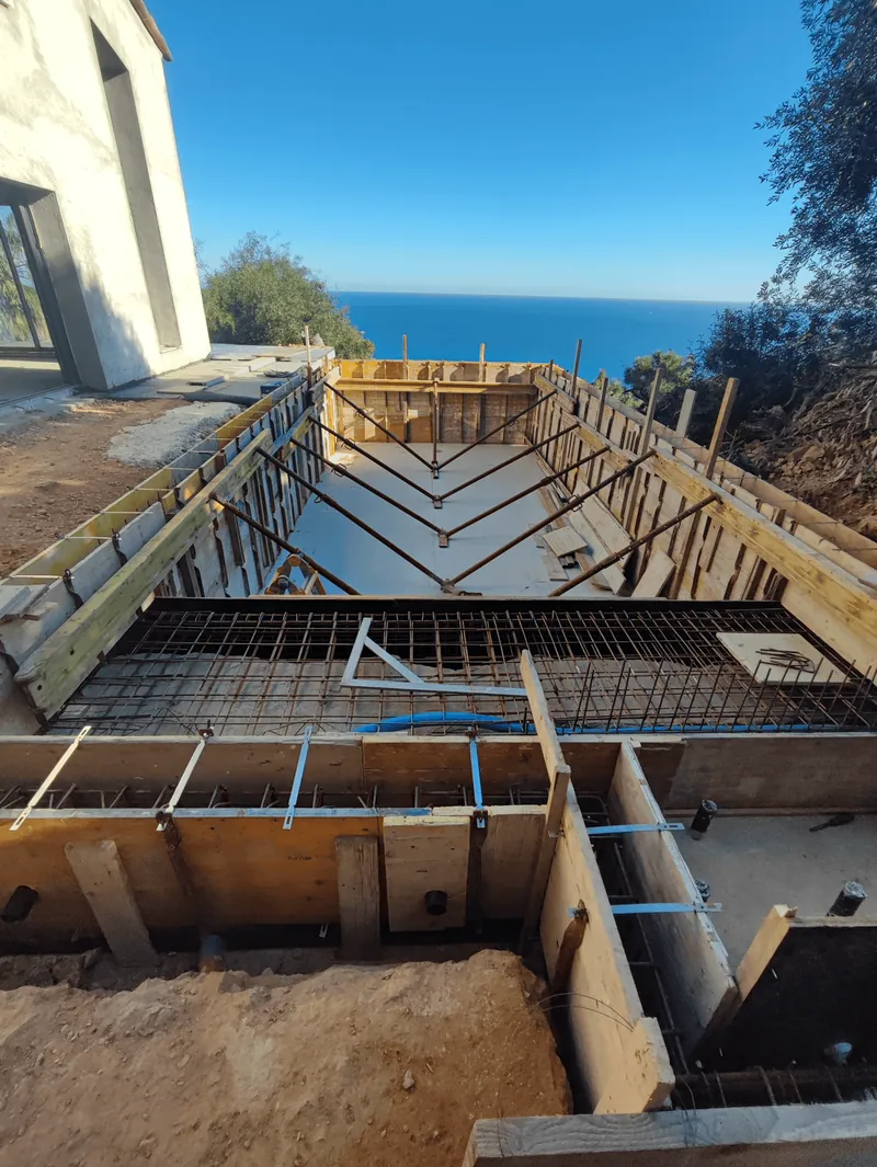 Bassin en construction avec coffrages, armatures et étais à côté d’une maison.